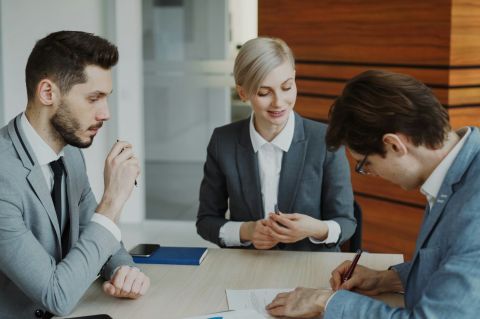Young businessman in suit signing contract with his colleague while corporate lawyer explaining details of partnership sitting in modern office indoors