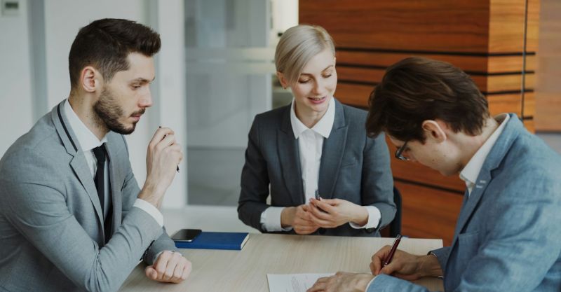 Young businessman in suit signing contract with his colleague while corporate lawyer explaining details of partnership sitting in modern office indoors